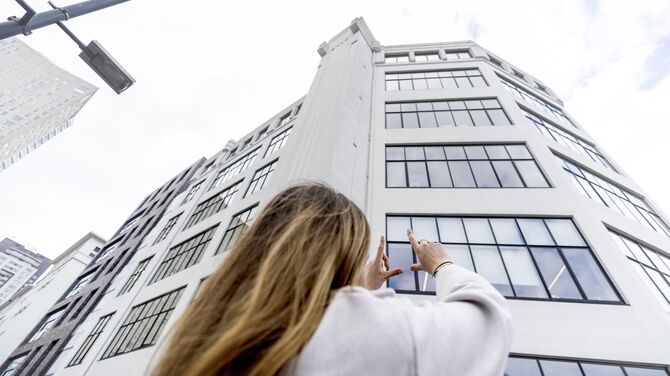 Mujer sosteniendo sus manos hacia un edificio blanco de mediana altura con muchas VENTANAS.