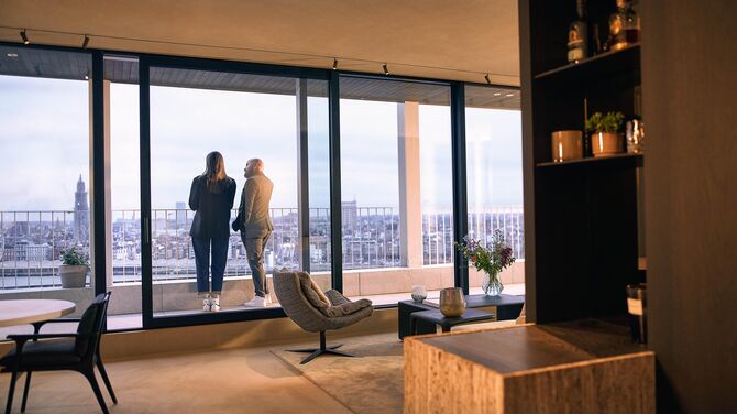 Alt: Vista interior de un moderno departamento con amplias puertas correderas y dos personas en el balcón.