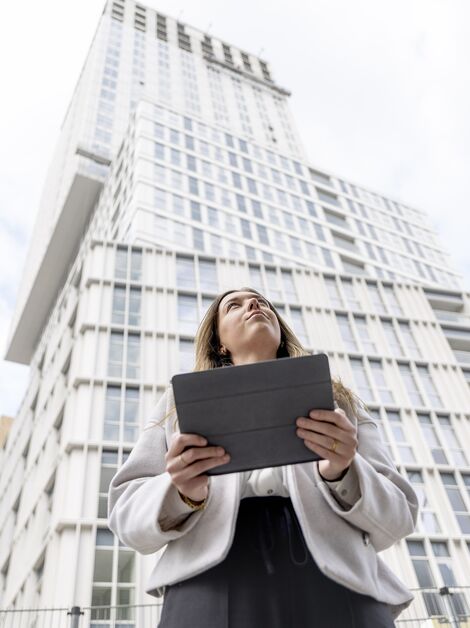 Mujer rubia sosteniendo un iPad y mirando hacia un edificio de apartamentos blanco.