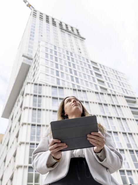 Mujer rubia sosteniendo un iPad y mirando hacia un edificio de apartamentos blanco.